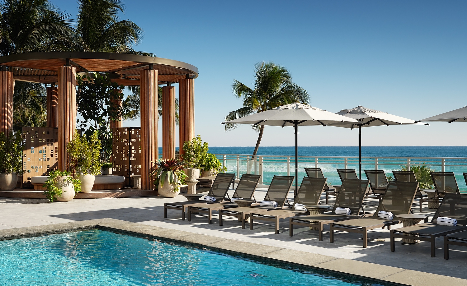 Lounge chairs with towels are arranged beside a blue swimming pool, on a sunny deck overlooking a vibrant ocean, with a wooden cabana.