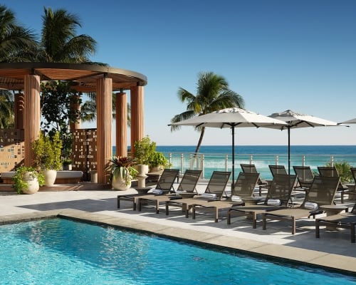 Lounge chairs with towels are arranged beside a blue swimming pool, on a sunny deck overlooking a vibrant ocean, with a wooden cabana.