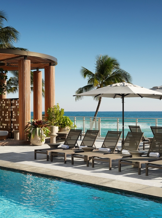 Lounge chairs with towels are arranged beside a blue swimming pool, on a sunny deck overlooking a vibrant ocean, with a wooden cabana.