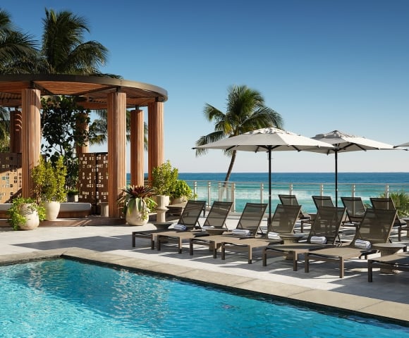 Lounge chairs with towels are arranged beside a blue swimming pool, on a sunny deck overlooking a vibrant ocean, with a wooden cabana.