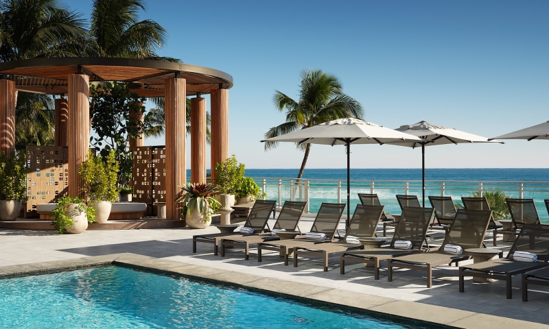 Lounge chairs with towels are arranged beside a blue swimming pool, on a sunny deck overlooking a vibrant ocean, with a wooden cabana.