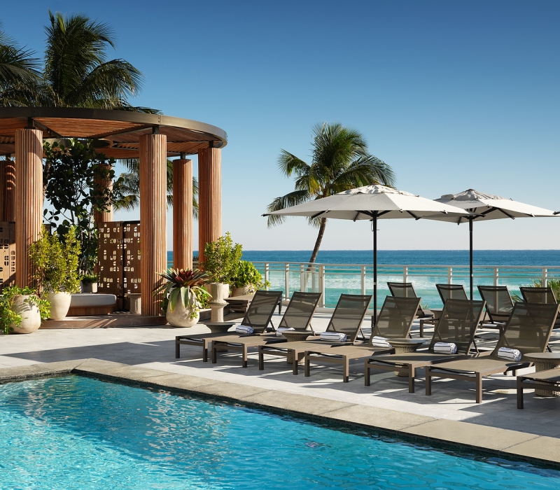 Lounge chairs with towels are arranged beside a blue swimming pool, on a sunny deck overlooking a vibrant ocean, with a wooden cabana.