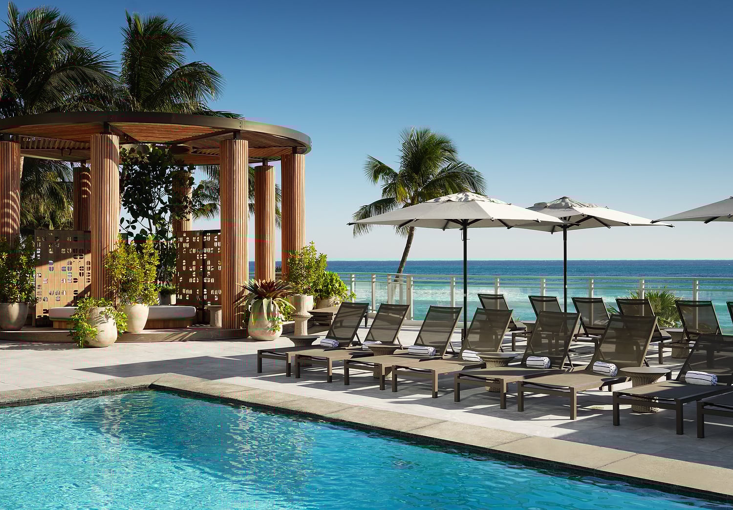 Lounge chairs with towels are arranged beside a blue swimming pool, on a sunny deck overlooking a vibrant ocean, with a wooden cabana.