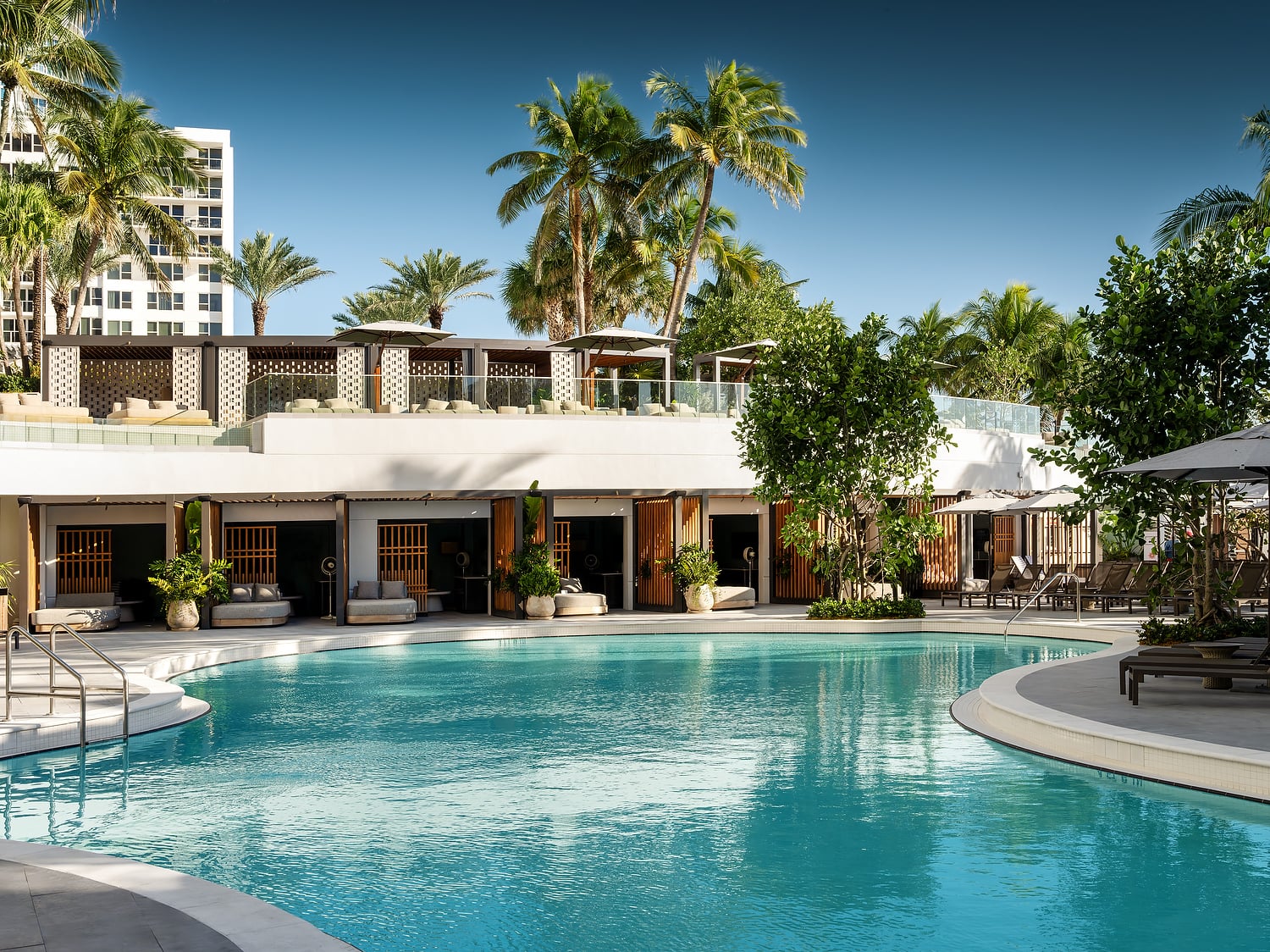 A resort swimming pool is surrounded by cabanas, loungers, and palm trees under a clear blue sky, with a tall white building in the background.