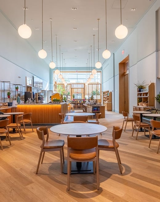 A modern cafe interior features light wood floors, numerous tables and chairs, and a long service counter. Spherical pendant lights hang from the high ceiling. Text: FRESH.