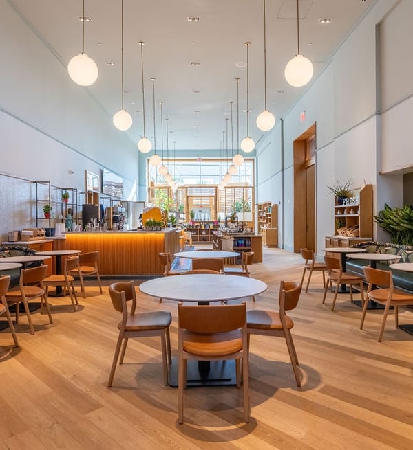 A modern cafe interior features light wood floors, numerous tables and chairs, and a long service counter. Spherical pendant lights hang from the high ceiling. Text: FRESH.