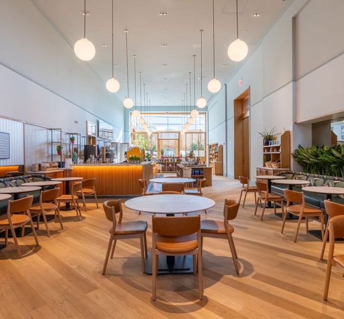 A modern cafe interior features light wood floors, numerous tables and chairs, and a long service counter. Spherical pendant lights hang from the high ceiling. Text: FRESH.