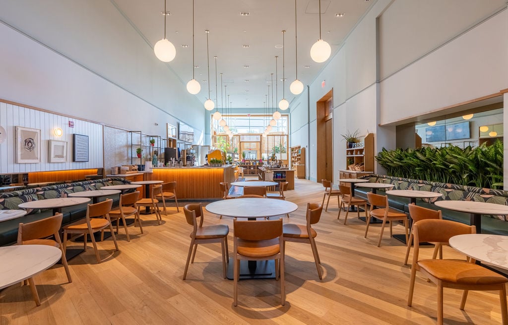 A modern cafe interior features light wood floors, numerous tables and chairs, and a long service counter. Spherical pendant lights hang from the high ceiling. Text: FRESH.