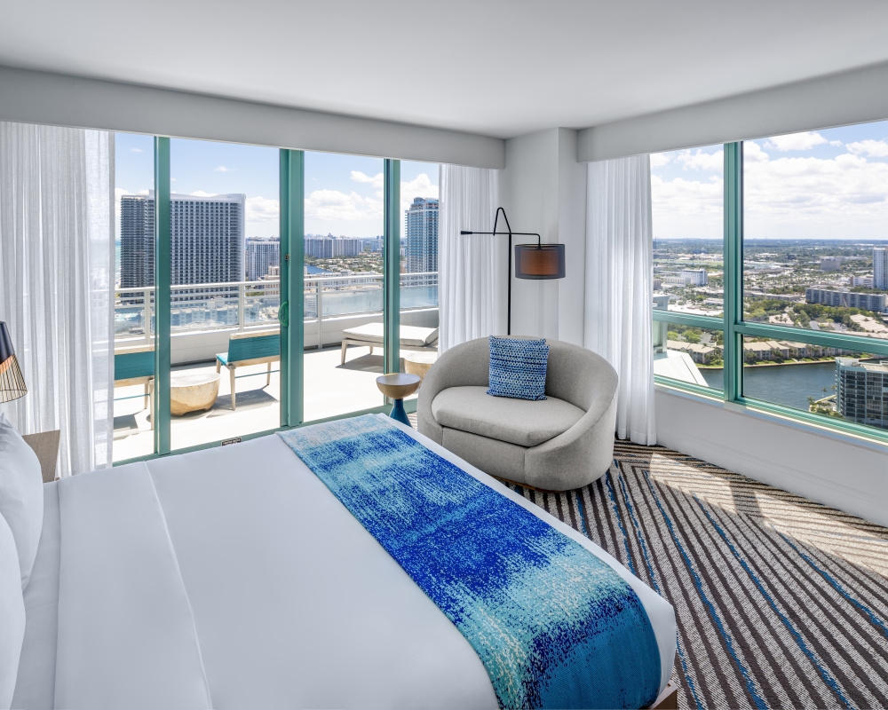 Modern hotel room features a white bed with blue runner, a grey armchair, and a balcony overlooking a city and waterway under a clear sky. Text: CAUTION WATCH YOUR STEP WET SURFACE SLIP HAZARD TO AVOID ACCIDENTS