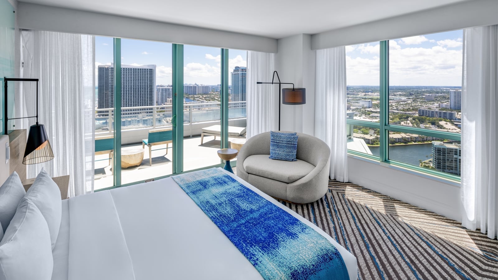 Modern hotel room features a white bed with blue runner, a grey armchair, and a balcony overlooking a city and waterway under a clear sky. Text: CAUTION WATCH YOUR STEP WET SURFACE SLIP HAZARD TO AVOID ACCIDENTS