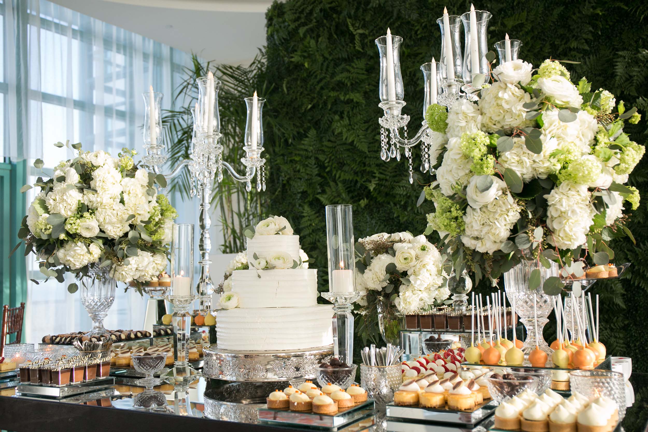 A lavish dessert table features a three-tiered white cake, various pastries, and lit crystal candelabras. White and green floral arrangements decorate it, set against sheer curtains and a plant wall.
