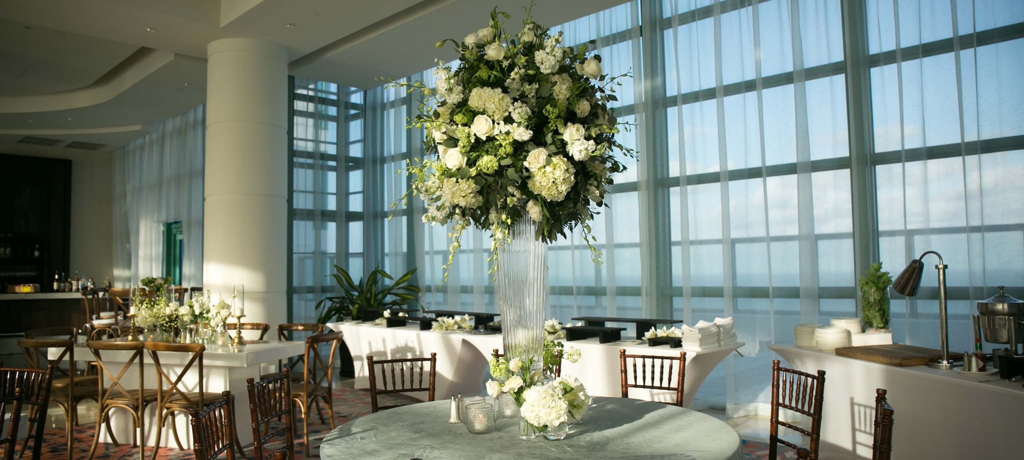 A towering white and green floral centerpiece is centered on a round table for an event setup in a modern venue with expansive windows against a bright sky.