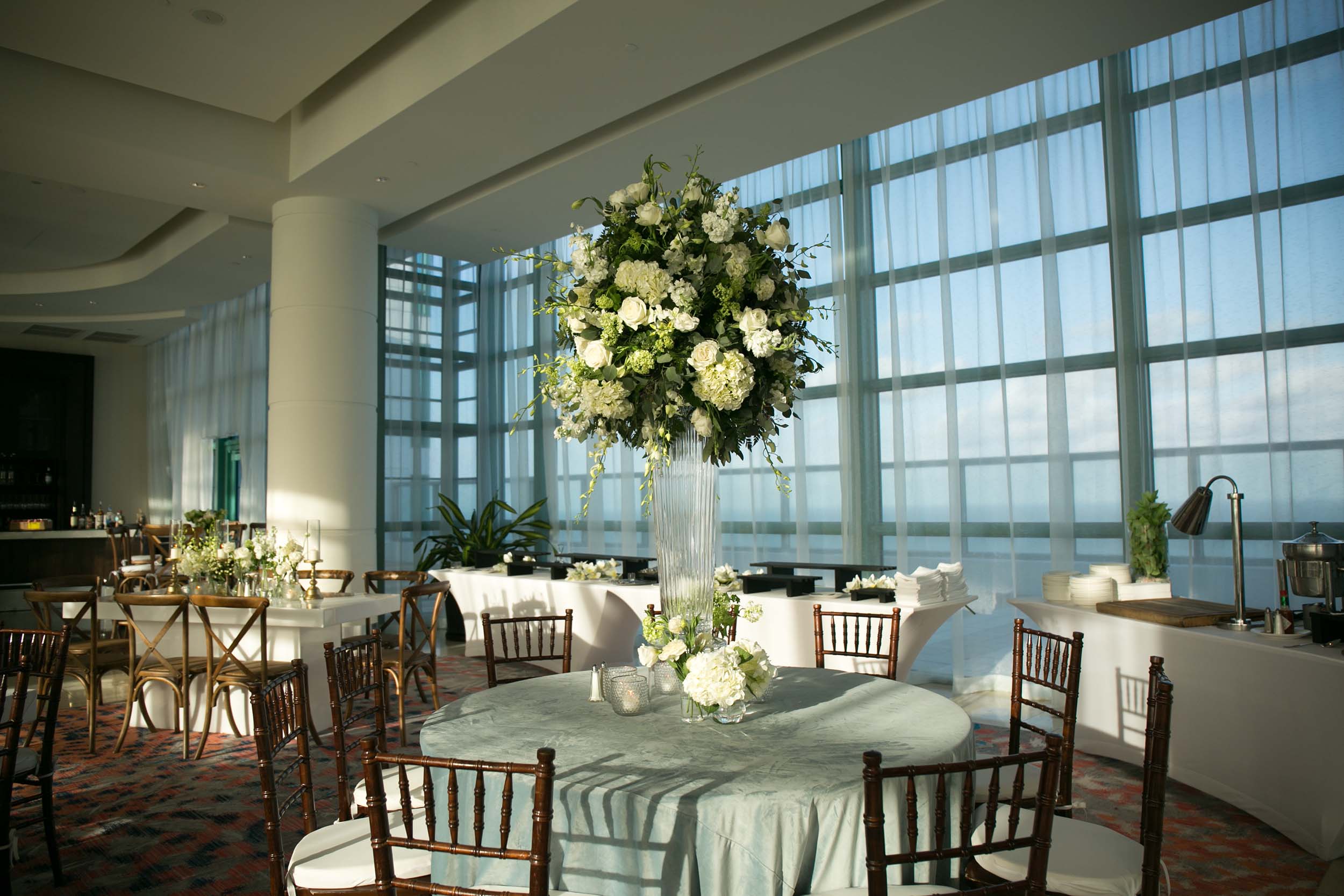 A towering white and green floral centerpiece is centered on a round table for an event setup in a modern venue with expansive windows against a bright sky.