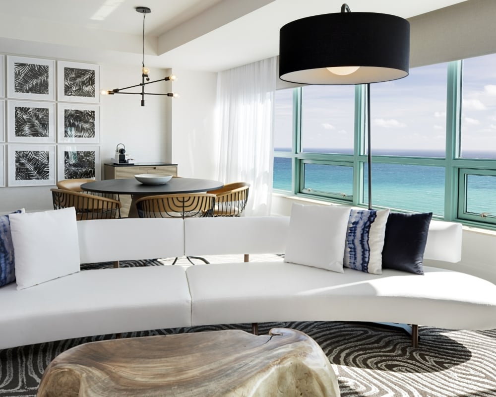 A contemporary white curved sofa with blue pillows sits on a striped rug, facing large ocean-view windows. A dining area and palm leaf art fill the background of the modern room.