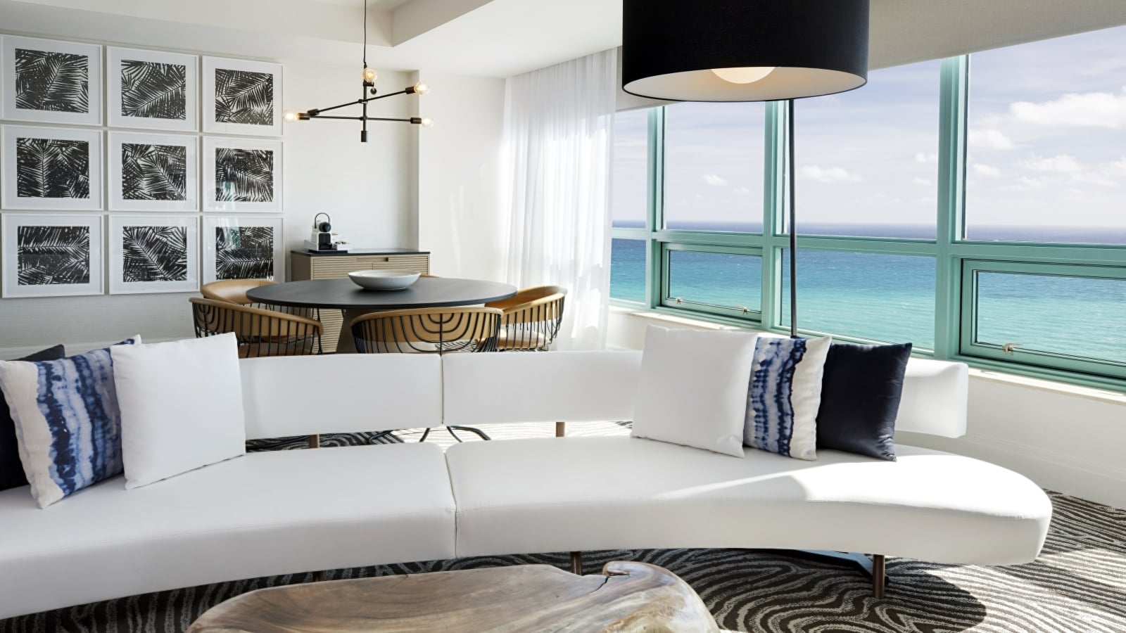 A contemporary white curved sofa with blue pillows sits on a striped rug, facing large ocean-view windows. A dining area and palm leaf art fill the background of the modern room.