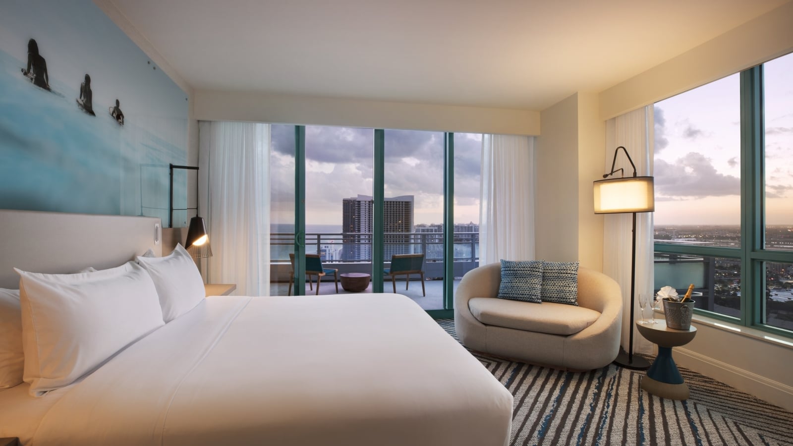 A modern hotel room, showcasing a large bed and curved chaise lounge, overlooks a city and ocean from a balcony at dusk. A blue surfer mural adorns the wall.