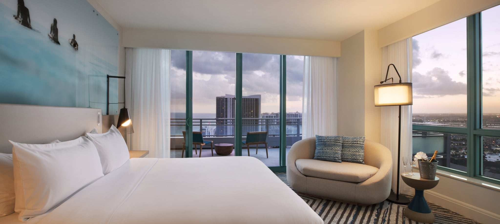 A modern hotel room, showcasing a large bed and curved chaise lounge, overlooks a city and ocean from a balcony at dusk. A blue surfer mural adorns the wall.
