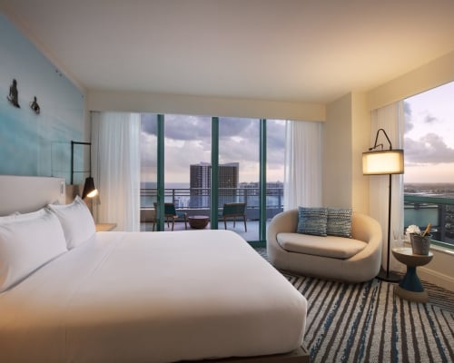 A modern hotel room, showcasing a large bed and curved chaise lounge, overlooks a city and ocean from a balcony at dusk. A blue surfer mural adorns the wall.