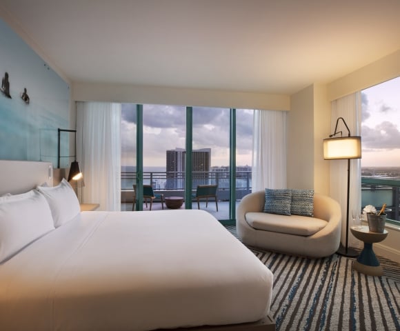 A modern hotel room, showcasing a large bed and curved chaise lounge, overlooks a city and ocean from a balcony at dusk. A blue surfer mural adorns the wall.