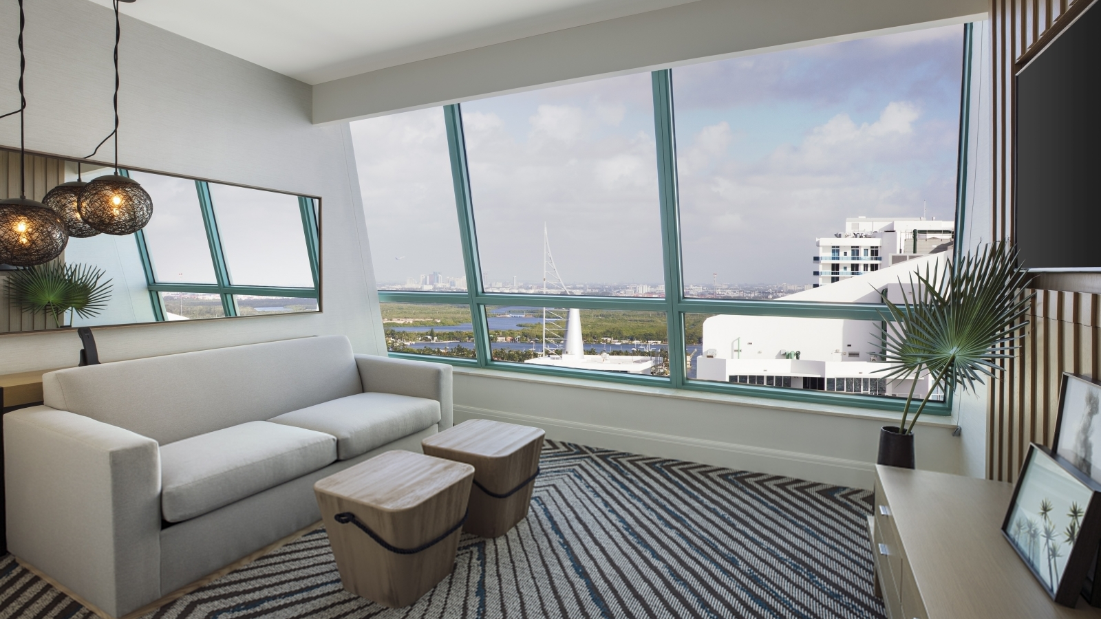 A modern living room features a light grey sofa, two wooden cube stools, and round pendant lights, with large angled windows revealing a city skyline over water.