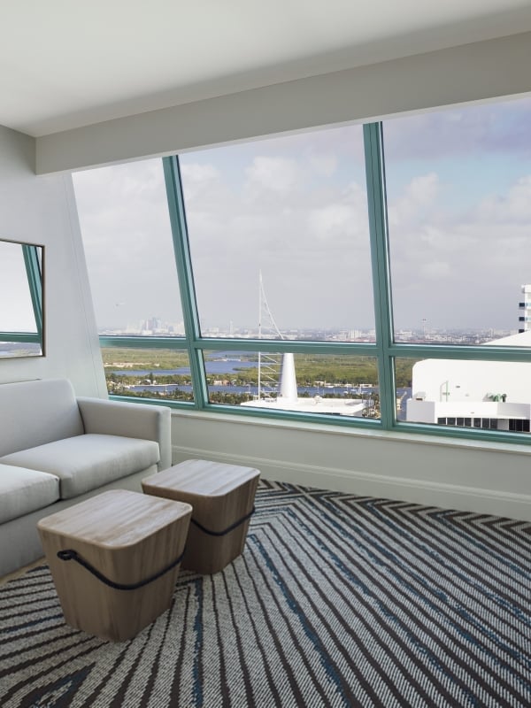 A modern living room features a light grey sofa, two wooden cube stools, and round pendant lights, with large angled windows revealing a city skyline over water.