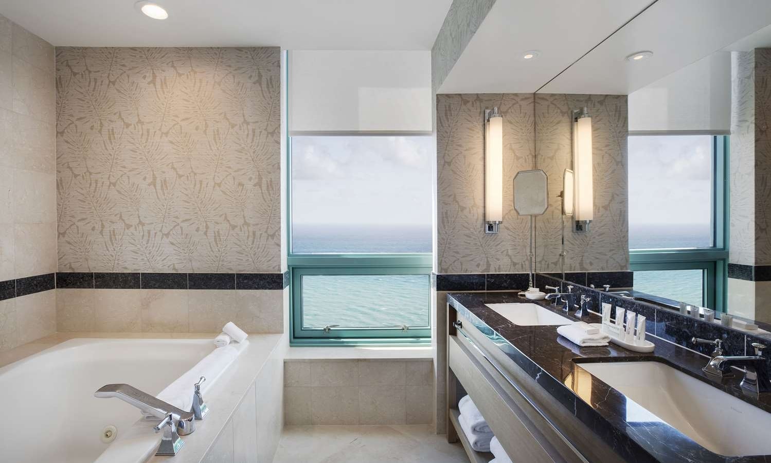 A modern, luxurious bathroom features a white soaking tub with chrome fixtures and a dark marble dual vanity. Large windows offer a stunning ocean view.