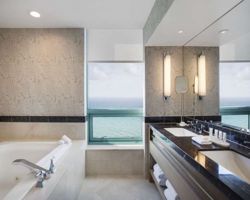 A modern, luxurious bathroom features a white soaking tub with chrome fixtures and a dark marble dual vanity. Large windows offer a stunning ocean view.