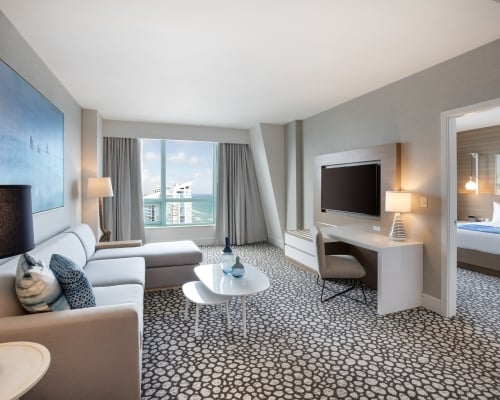 A modern hotel suite living area is furnished with an L-shaped sofa, nested white tables, and patterned carpet. A large window offers ocean views; an adjoining bedroom is visible.