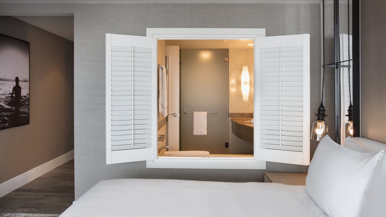Hotel guestroom featuring a white-bedding setup with open white window shutters above the headboard that look directly into an en-suite bathroom with a lighted vanity mirror.