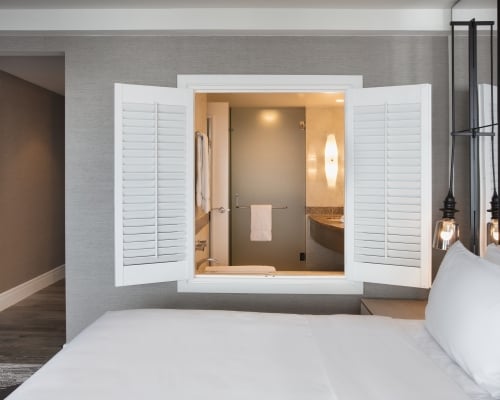 Hotel guestroom featuring a white-bedding setup with open white window shutters above the headboard that look directly into an en-suite bathroom with a lighted vanity mirror.