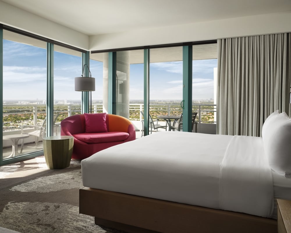 Corner king accommodation featuring white linens, a large red lounge chair, and floor to ceiling windows that look onto the patio and surrounding area.