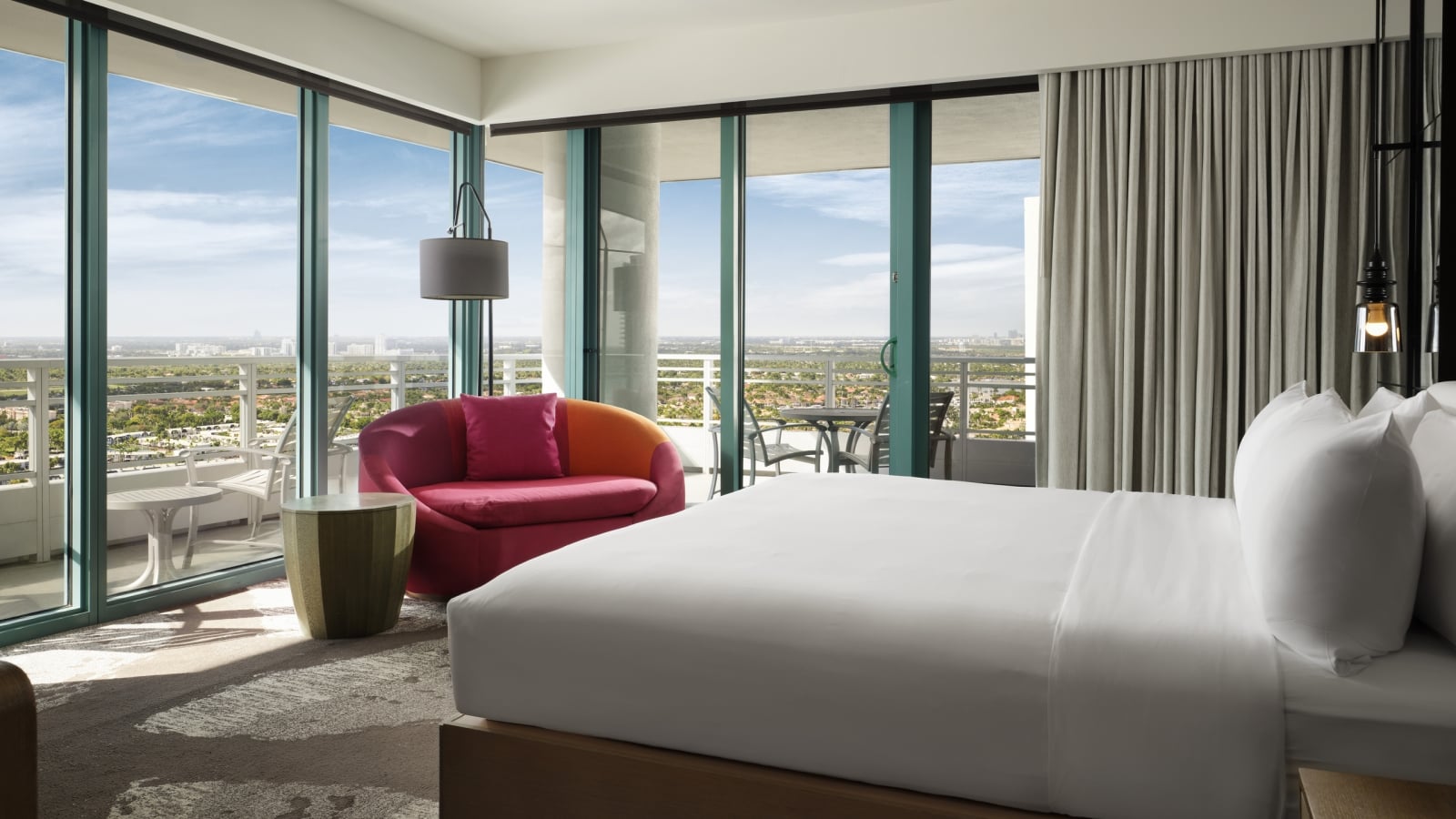 Corner king accommodation featuring white linens, a large red lounge chair, and floor to ceiling windows that look onto the patio and surrounding area.
