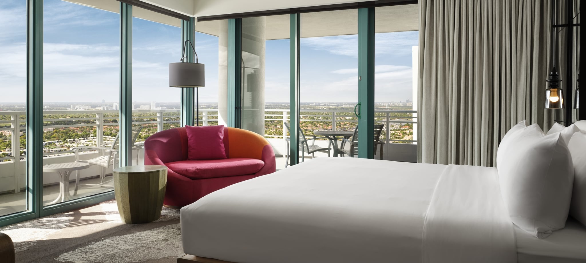 Corner king accommodation featuring white linens, a large red lounge chair, and floor to ceiling windows that look onto the patio and surrounding area.