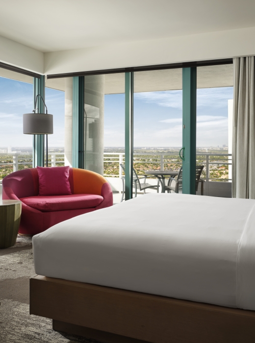 Corner king accommodation featuring white linens, a large red lounge chair, and floor to ceiling windows that look onto the patio and surrounding area.