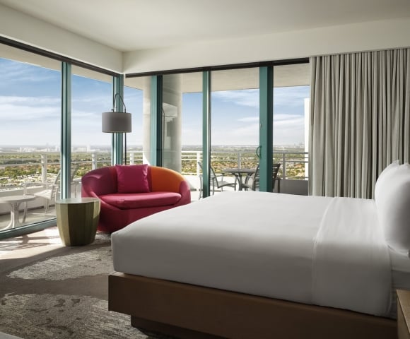 Corner king accommodation featuring white linens, a large red lounge chair, and floor to ceiling windows that look onto the patio and surrounding area.