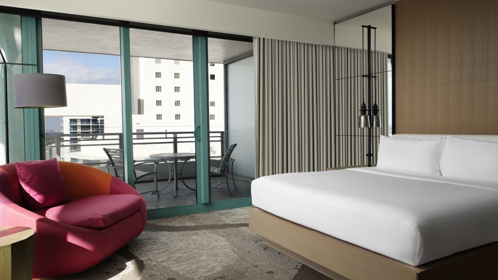 Corner king accommodation featuring white linens, a large red lounge chair, and floor to ceiling windows that look onto the patio and surrounding area.