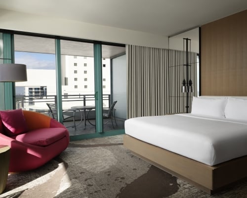 Corner king accommodation featuring white linens, a large red lounge chair, and floor to ceiling windows that look onto the patio and surrounding area.