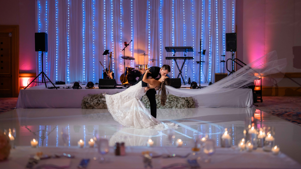 A groom dips and kisses his bride, her veil flowing, on a reflective white dance floor in front of a stage with blue string lights and band equipment. Blurred candles are in the foreground.