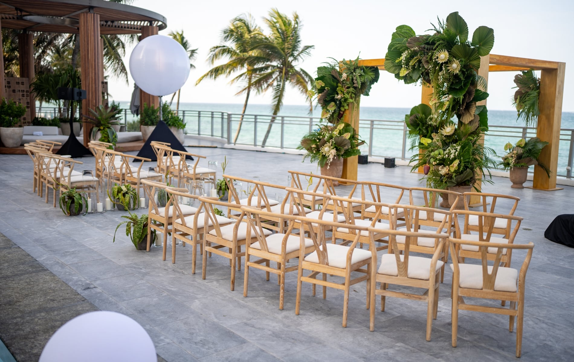An elegant pergola decorated with local plants is positioned center amongst rows of seating, setup for an outdoor gathering