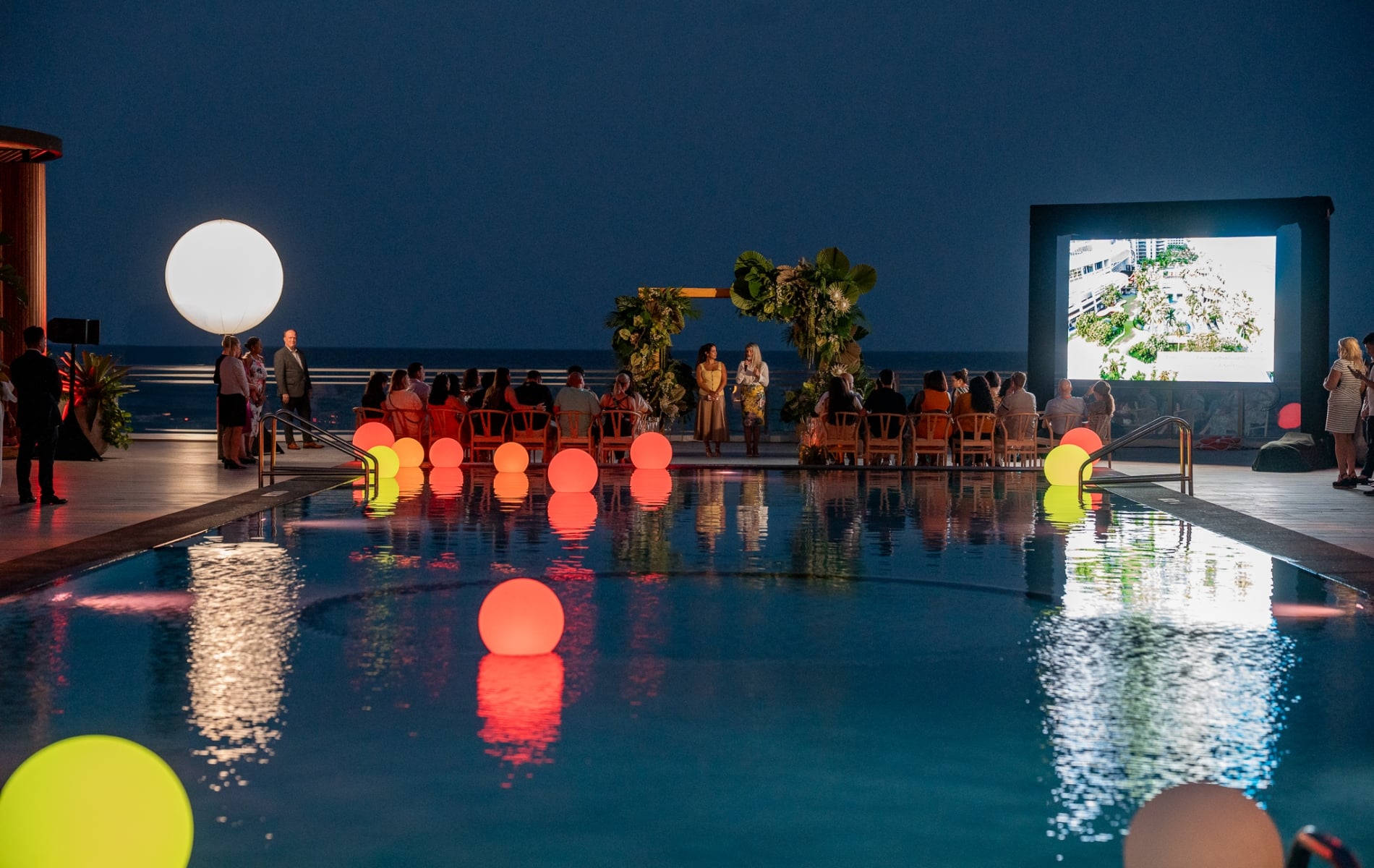 An outdoor gathering by the pool at night, featuring a fauna-centric pergola centered amongst rows of seating, and a large projector screen, positioned right.