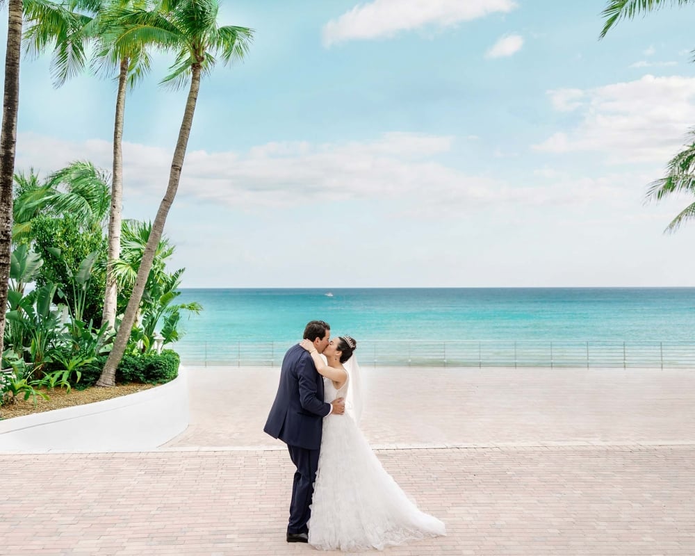 A bride and groom kiss on a brick walkway flanked by palm trees and lush greenery, with the turquoise ocean and a railing in the background.
