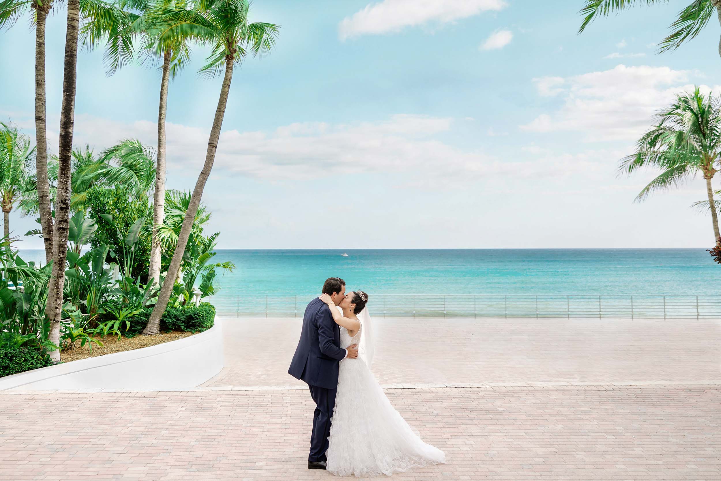 A bride and groom kiss on a brick walkway flanked by palm trees and lush greenery, with the turquoise ocean and a railing in the background.