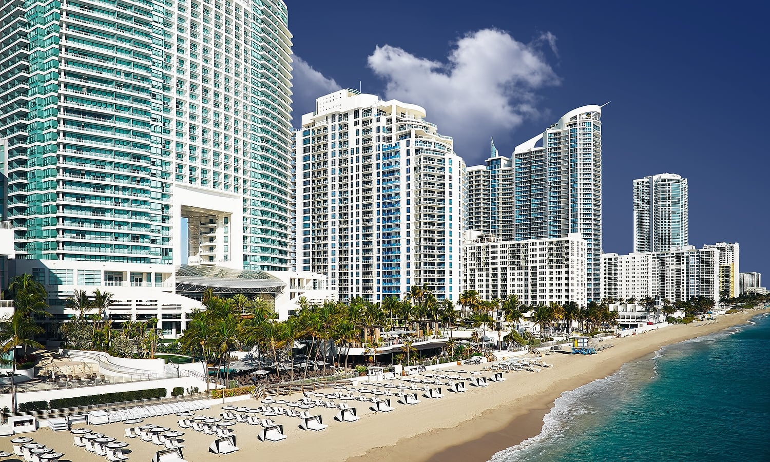 View of the beach featuring rows of lounge chairs and cabanas on a clear blue day