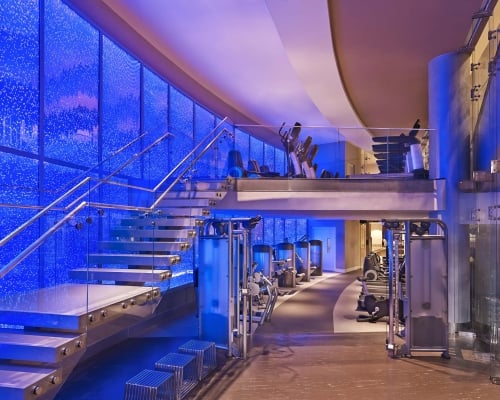A modern, multi-level gym displays Life Fitness equipment. It features a floating glass staircase ascending against a vibrant blue bubble wall.