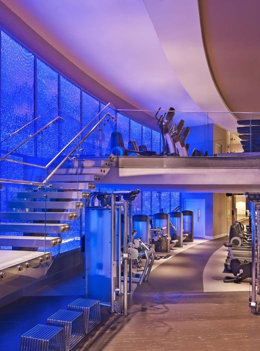 A modern, multi-level gym displays Life Fitness equipment. It features a floating glass staircase ascending against a vibrant blue bubble wall.