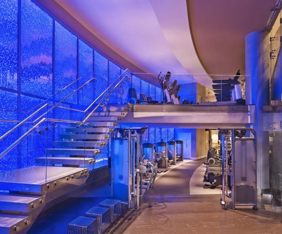 A modern, multi-level gym displays Life Fitness equipment. It features a floating glass staircase ascending against a vibrant blue bubble wall.