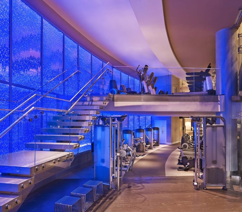 A modern, multi-level gym displays Life Fitness equipment. It features a floating glass staircase ascending against a vibrant blue bubble wall.
