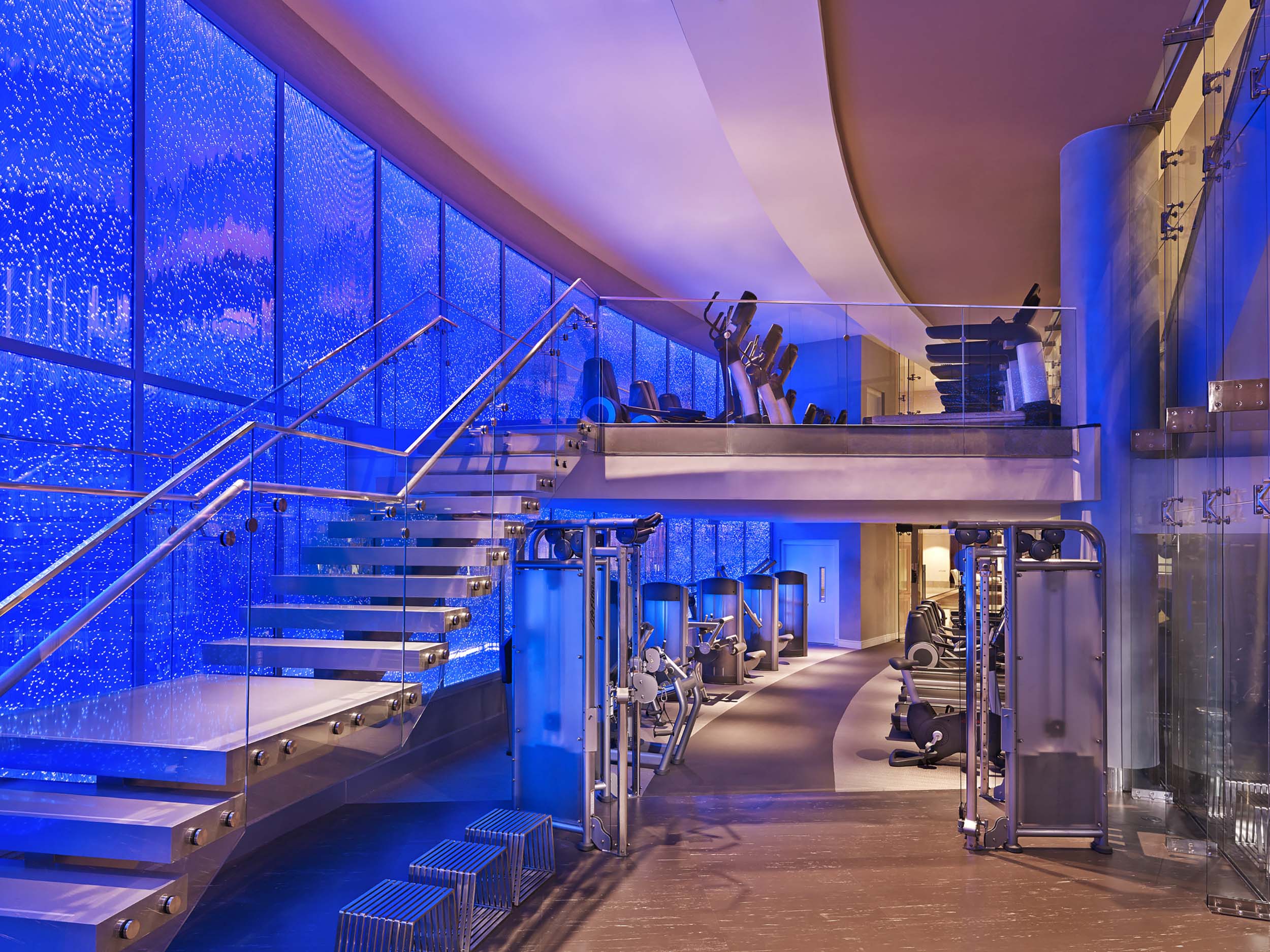 A modern, multi-level gym displays Life Fitness equipment. It features a floating glass staircase ascending against a vibrant blue bubble wall.