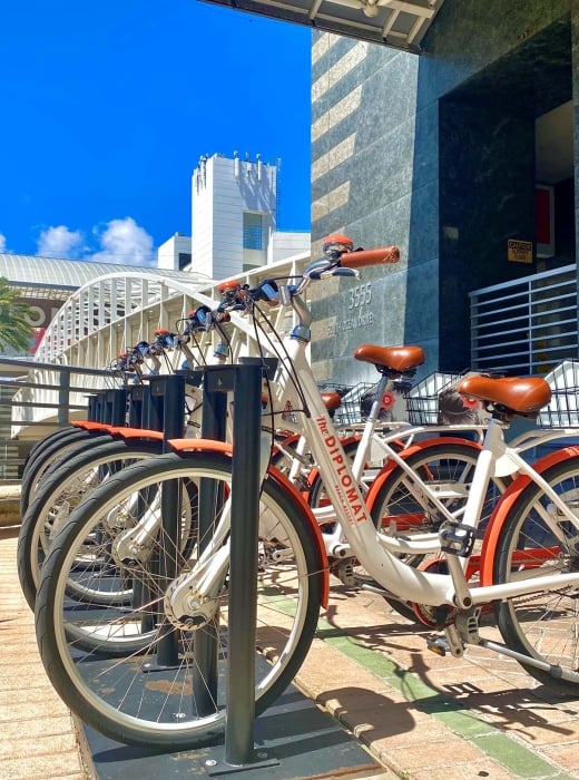 A row of white and orange "The DIPLOMAT BEACH RESORT" bicycles are parked on a paved plaza next to a dark building. Text: 3555 SOUTH OCEAN DRIVE, CAUTION SLIPPERY FLOOR.