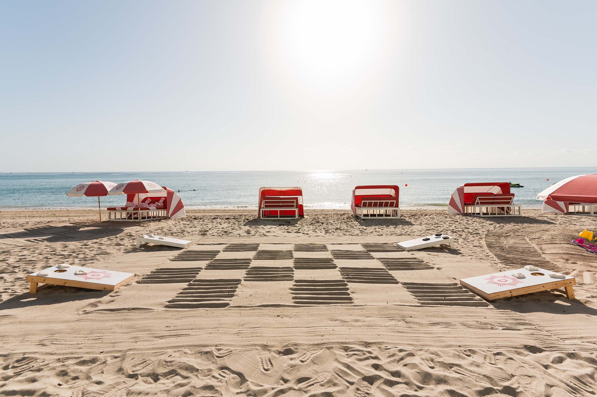 Two cornhole boards, each displaying a 'D' sunburst logo, are set on a sandy beach by a checkerboard pattern. Red-and-white beach chairs and umbrellas line the sunny ocean shore.